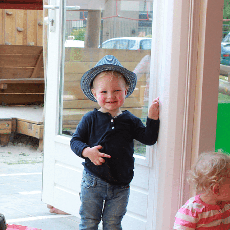 Ett litet barn iklädd blå skjorta och randig hatt ler och lutar sig mot en dörrkarm med Klämskydd för innerdörr, 195cm Vit. Ett annat barn i rosa outfit sitter i närheten.