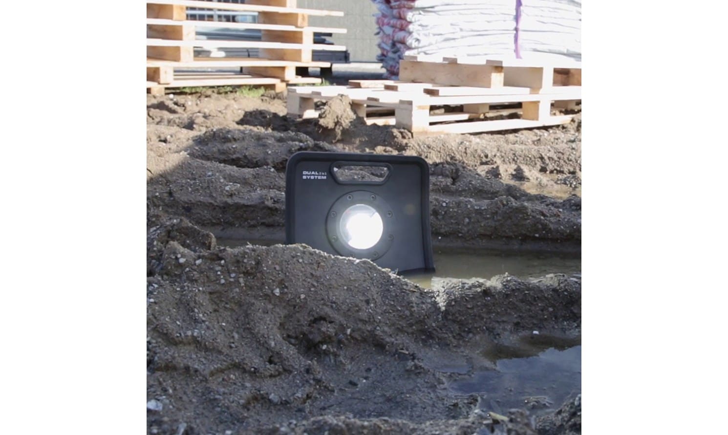 En bärbar Arbetslampa NOVA 3K C+R COB LED är delvis nedsänkt i en lerig pöl på en byggarbetsplats, omgiven av smuts, träpallar och påsar.