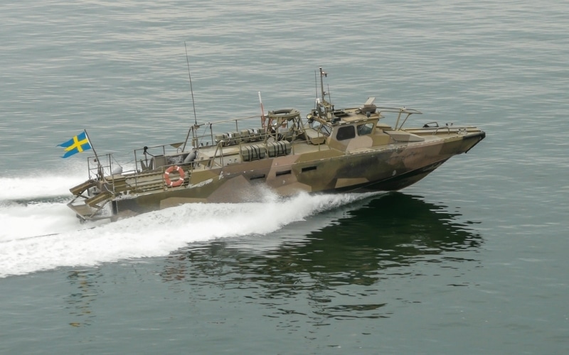 En militär patrullbåt med svensk flagg rör sig i hög fart över lugnt vatten och skapar ett vitt kölvatten efter sig. Båten är kamouflerad och försedd med antenner och diverse utrustning.