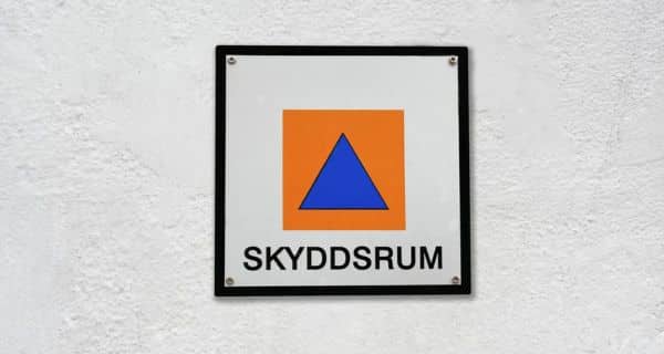 En fyrkantig skylt med en blå triangel inuti en orange fyrkant ovanför ordet "SKYDDSRUM" på en vit vägg, som anger ett svenskt skyddsrum.